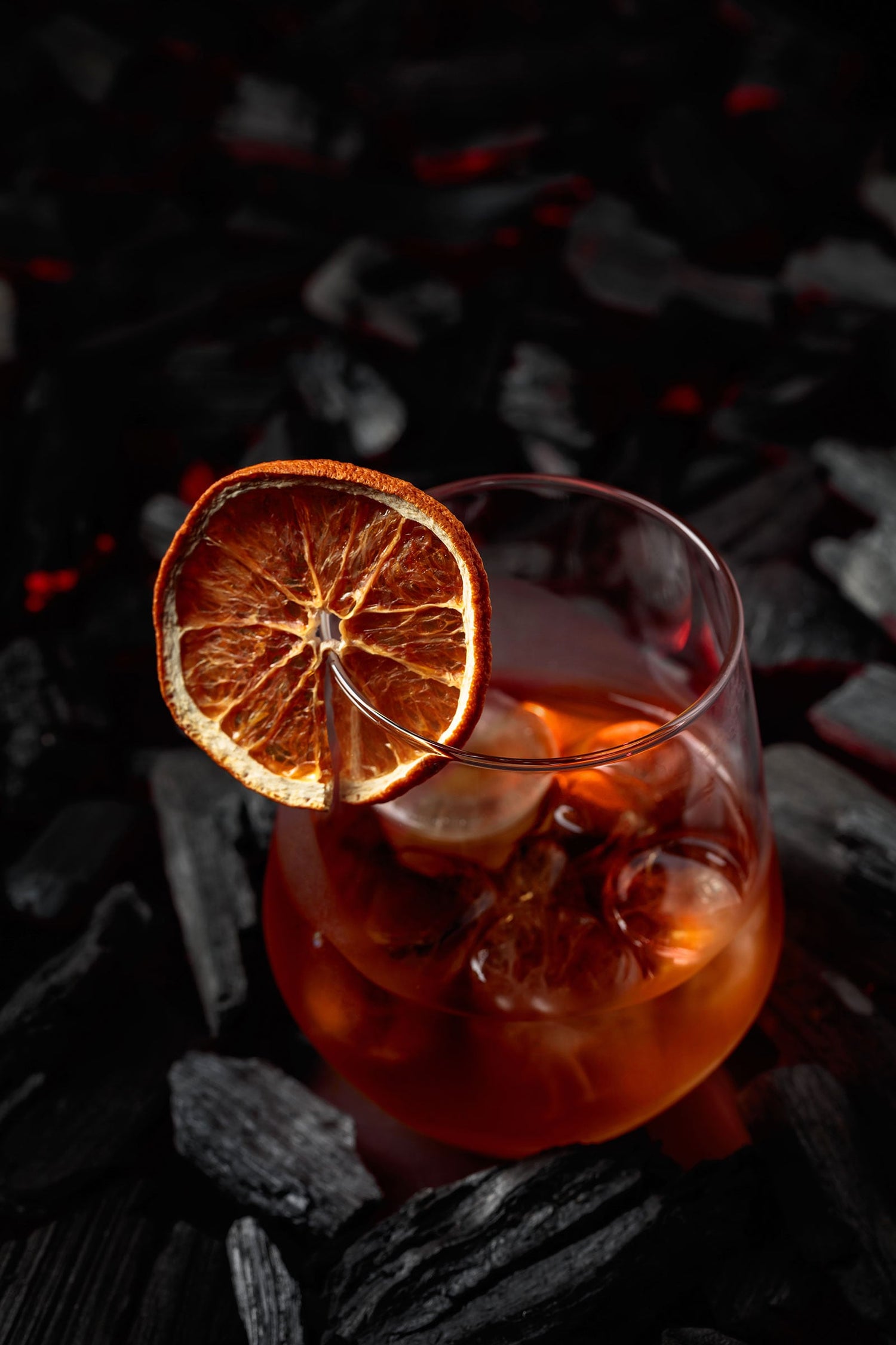 red cocktail drink in a stemless glass with a dehydrated fruit garnish, against a black background
